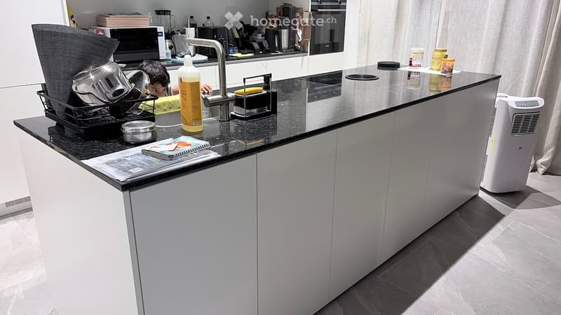 Modern kitchen with white cabinets, black countertop, and various appliances including a dish rack, oven, and air conditioner. A child is partially visible behind the counter.