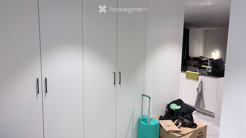 A modern, minimalist room with white built-in wardrobes, a small kitchen area with a sink, and a suitcase and backpack placed on the floor.