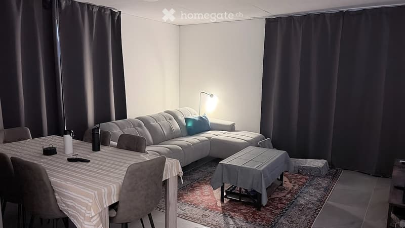 Modern living room with a grey sectional sofa, coffee table, dining table with chairs, and a floor lamp. The room features large windows with dark curtains and a patterned rug.
