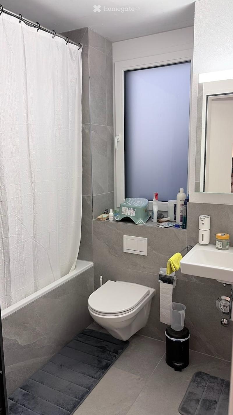 Modern bathroom with grey tiles, a toilet, a sink, a bathtub, a shower curtain, a mirror, and a small cabinet with toiletries.