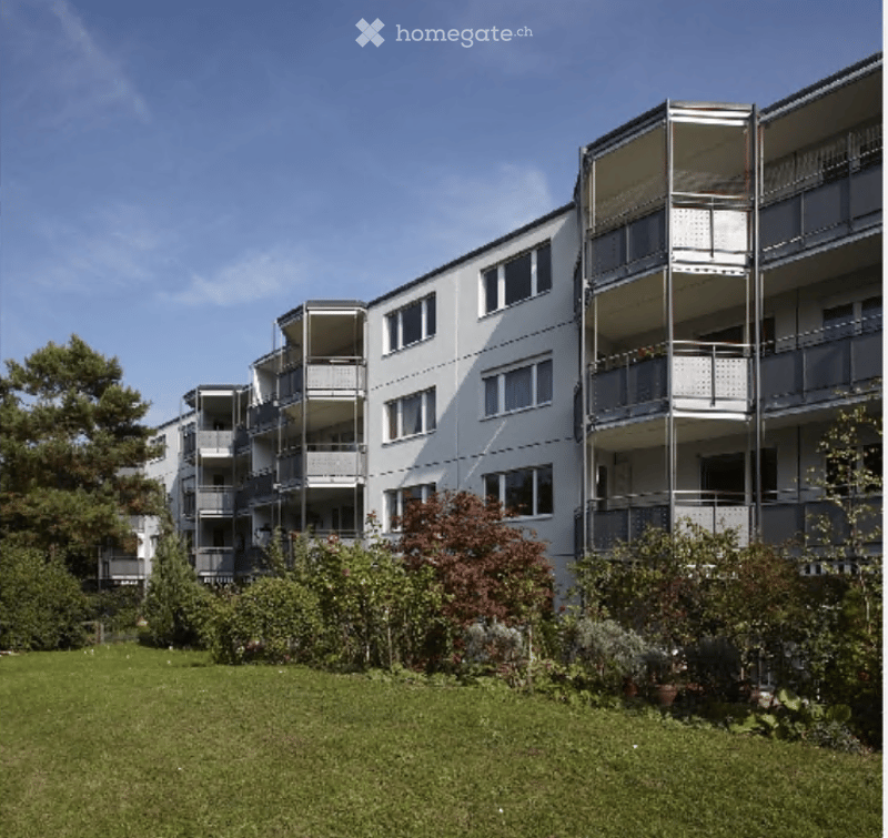 Modern multi-story apartment building with white facade, multiple balconies, and surrounding greenery.