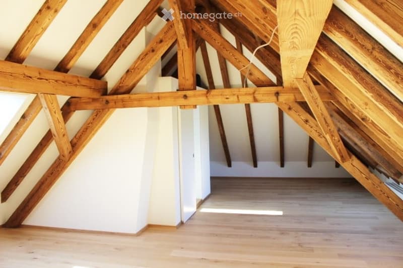 An attic space with exposed wooden beams and a wooden floor, featuring white walls and a small built-in closet.