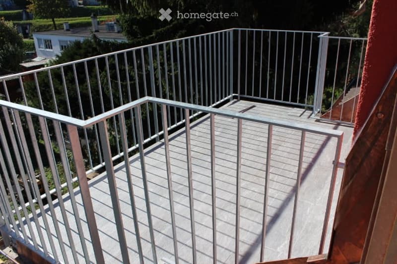 Balcony with metal railing and wooden decking, overlooking a residential area with trees and houses.