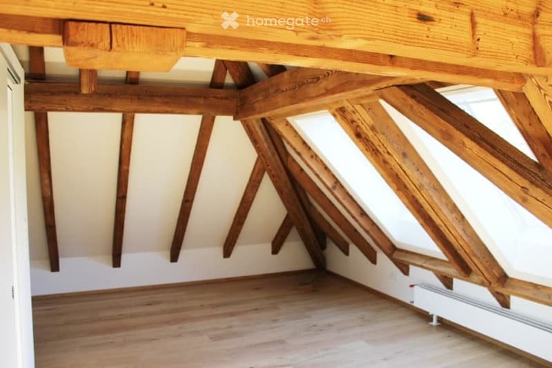 An empty attic space with exposed wooden beams and a wooden floor, featuring white walls and a small window letting in natural light.