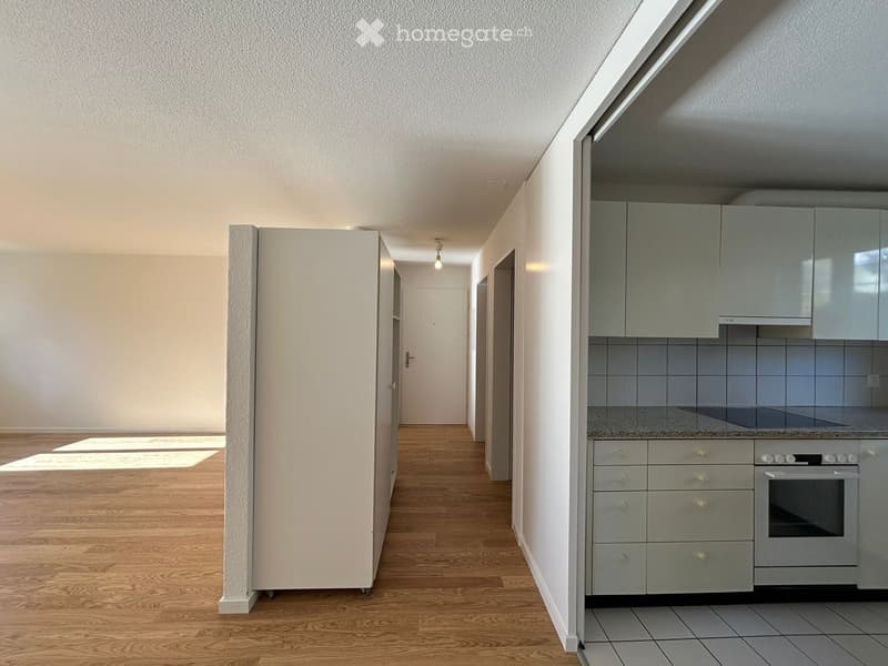 A bright, empty hallway with wooden flooring and white walls, leading to a modern kitchen area with grey cabinets and stainless steel appliances.