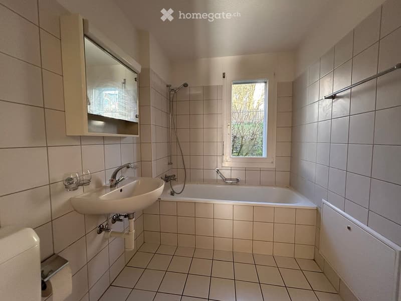 Bathroom with tiled walls and floor, a sink, bathtub, mirror, and a small window with a view of greenery.