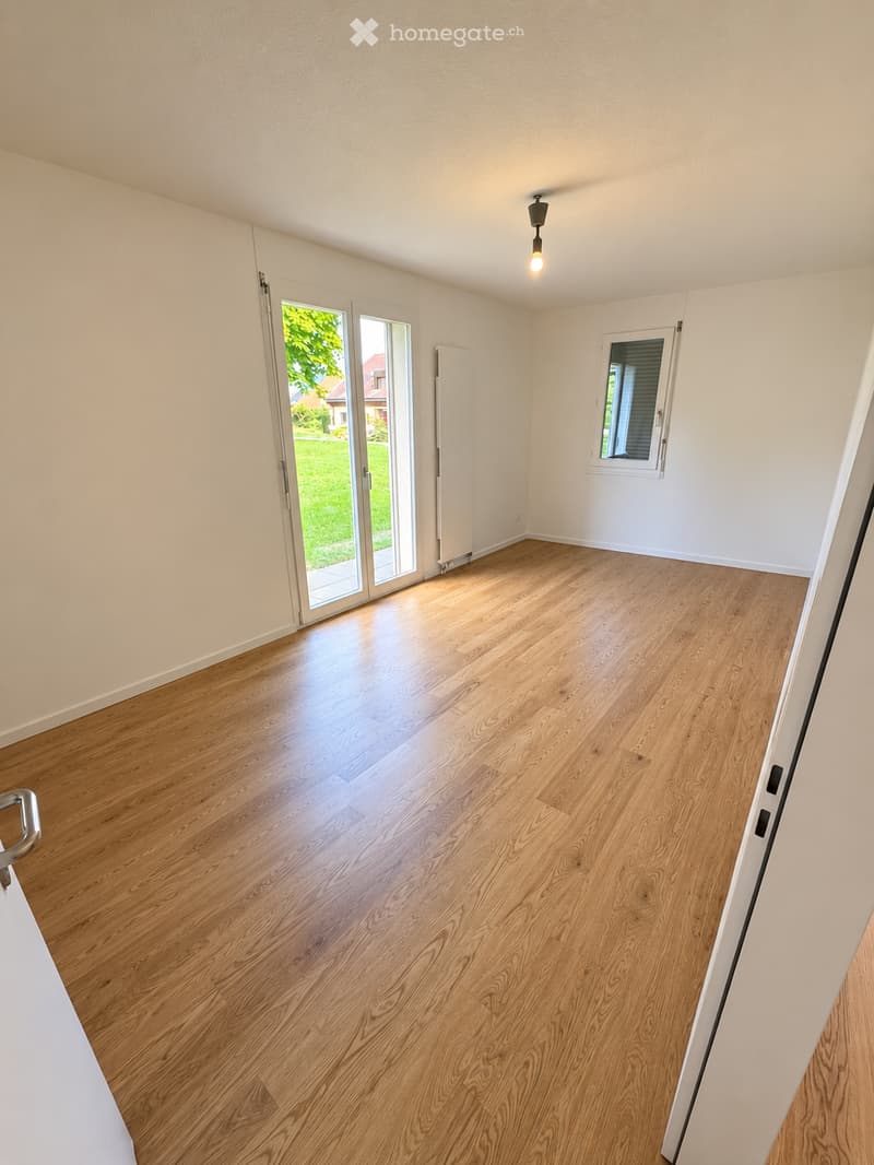Empty room with light wooden flooring, white walls, and large sliding glass doors leading to a green outdoor area.