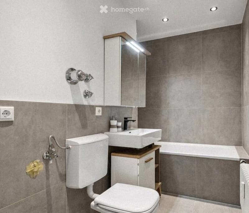 Modern bathroom with a white toilet, sink, bathtub, and mirrored cabinet. Grey tiled walls and floor, with a small wooden cabinet under the sink.