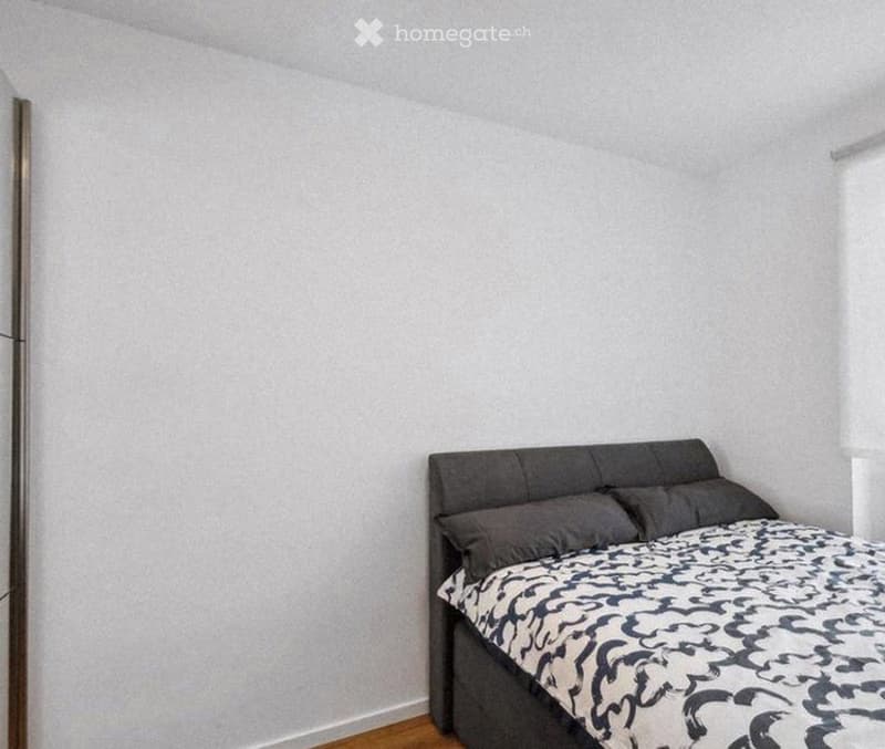 A small bedroom with a single bed, grey headboard, and patterned bedspread. The room has light grey walls and wooden flooring.