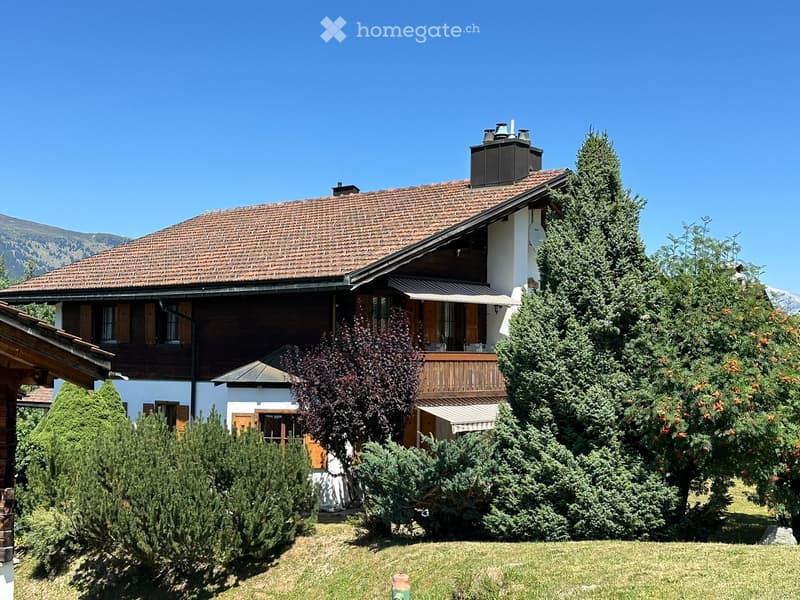 Two-story house with a tiled roof, white walls, wooden shutters, and a balcony. Surrounded by well-maintained greenery and trees.