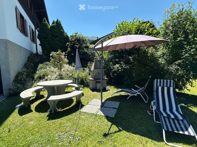 Garden area with a round stone table, benches, two lounge chairs, a large umbrella, and a stone fireplace.
