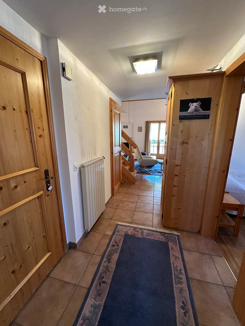 A narrow hallway with tiled flooring, wooden doors, a radiator, and a blue runner rug. A staircase leads to another room with visible furniture.