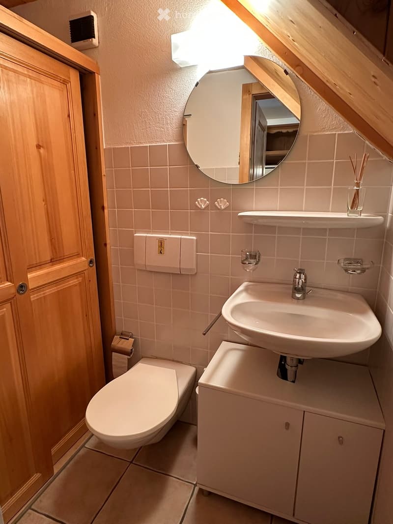 Cozy attic bathroom with a sink, toilet, round mirror, wooden cabinet, and decorative shelf with a vase.