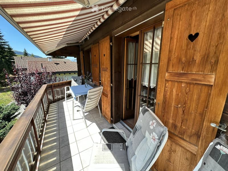 Balcony with wooden railings, tiled floor, and outdoor furniture including a table, chairs, and an umbrella. The balcony has a view of a garden and mountains.