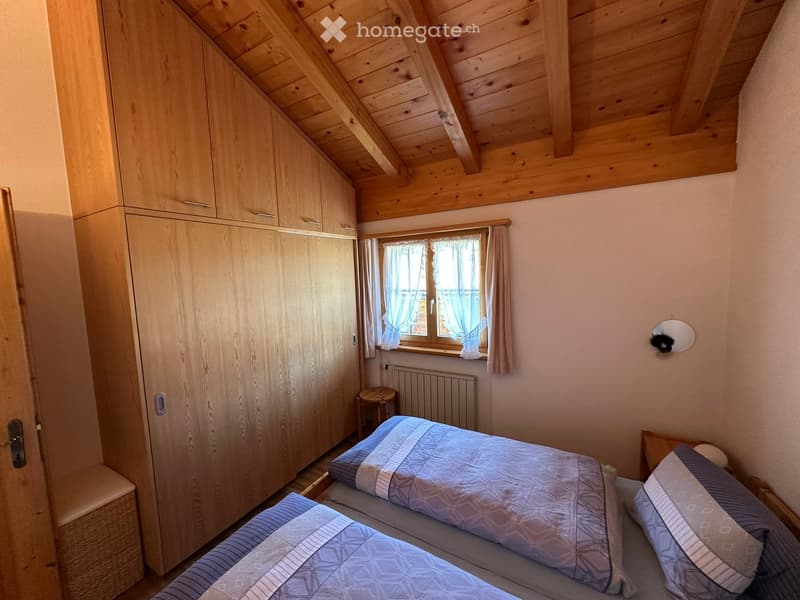 Cozy bedroom with a double bed, wooden wardrobe, small stool, radiator, and a window with curtains.