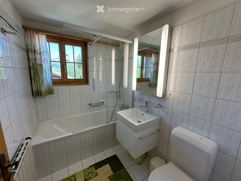Modern bathroom with a bathtub, sink, toilet, mirror, and a window with a curtain. The room features tiled walls and a green bathroom rug.