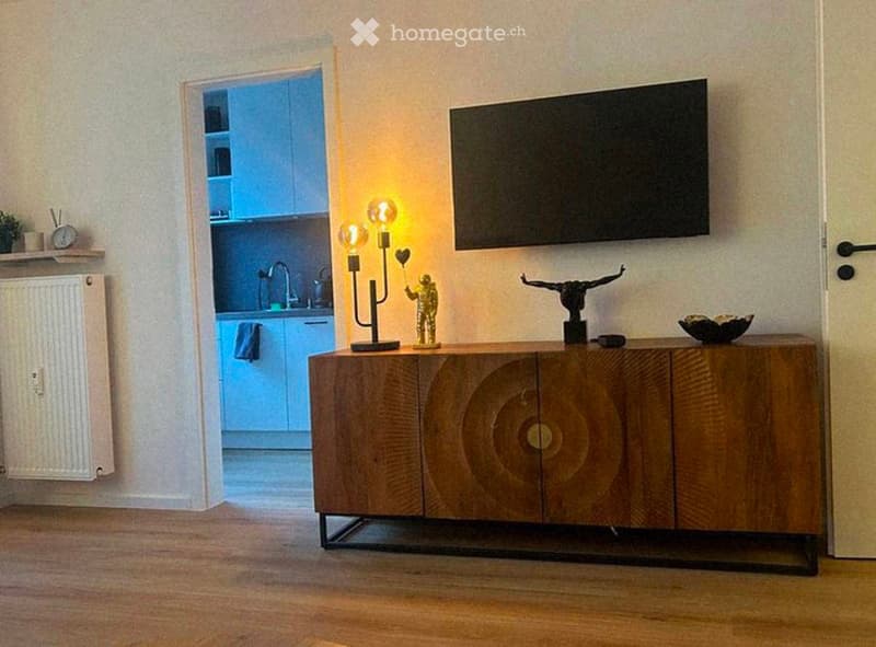 A modern living room with a wooden cabinet, table lamp, decorative statues, and a view into a small kitchen area with a sink and faucet.