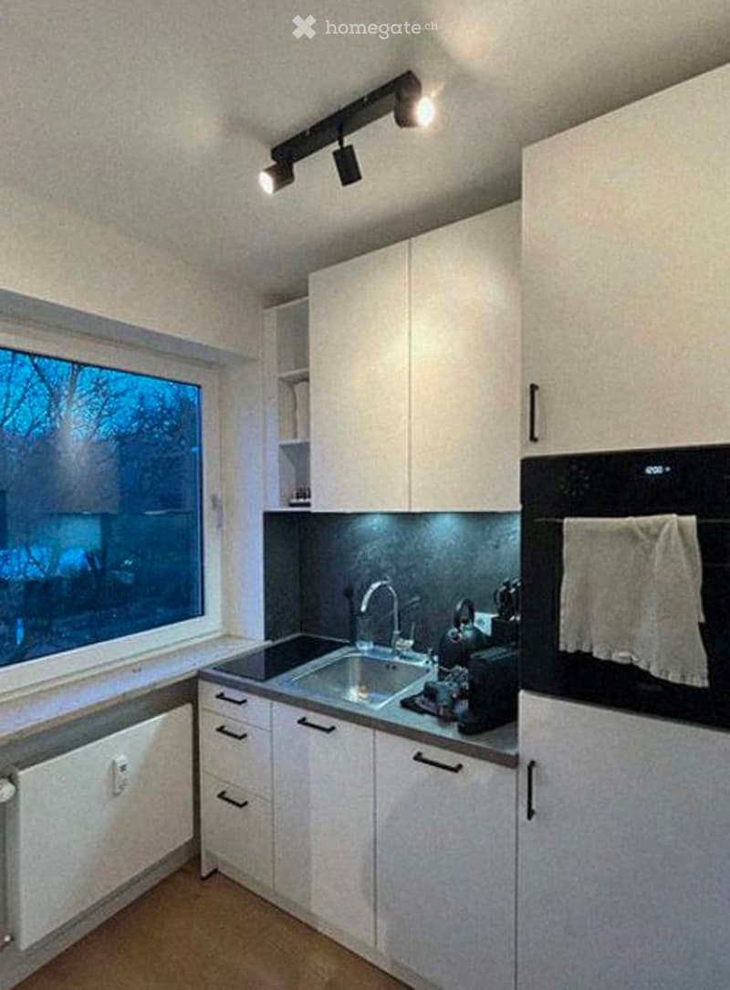 Modern kitchen with white cabinets, stainless steel sink, oven, and coffee maker on the counter. A large window provides natural light.