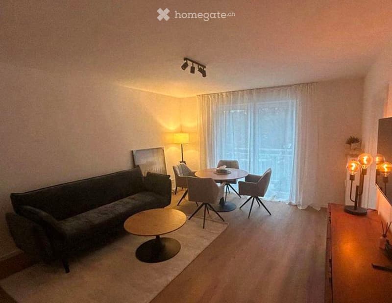 Modern living room with a grey sofa, coffee table, dining table with chairs, and decorative lamps. Large window with sheer curtains and a view of the balcony.