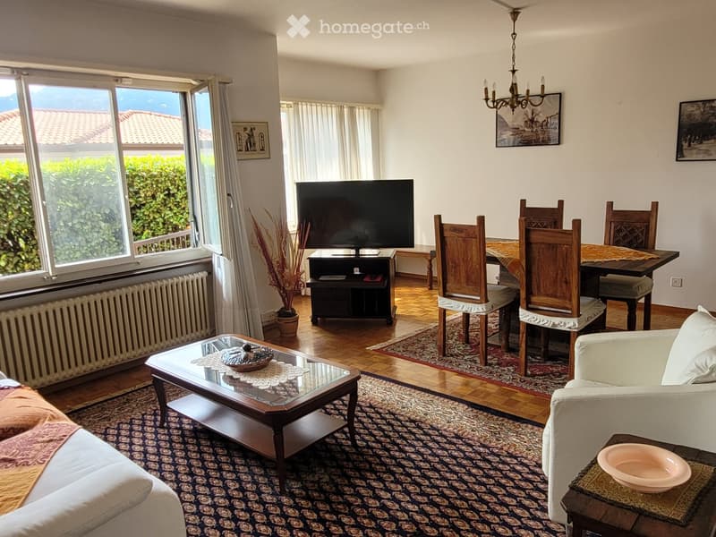 Spacious living room with large windows overlooking a balcony or terrace, hardwood floors, a wooden dining table with chairs, a TV stand, and a patterned area rug. The room has a cozy and rustic feel with wooden furniture and decor elements.