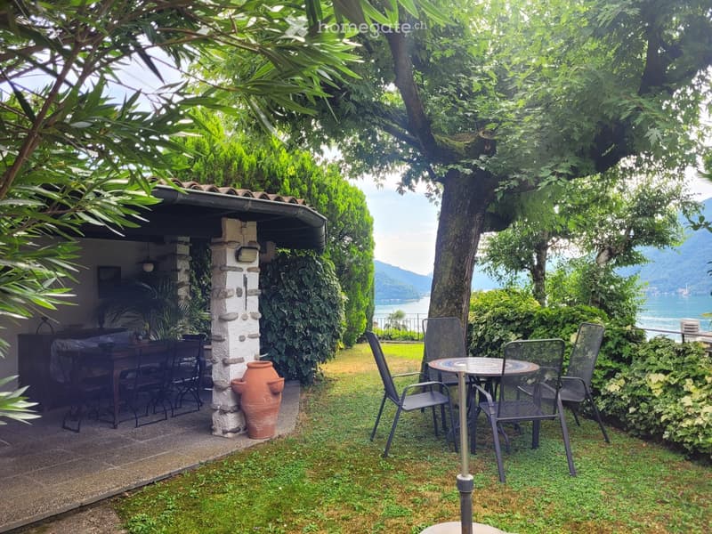 Cozy outdoor seating area with a stone structure, surrounded by lush greenery and trees. There is a table and chairs set up, with a view of the lake or body of water in the background.