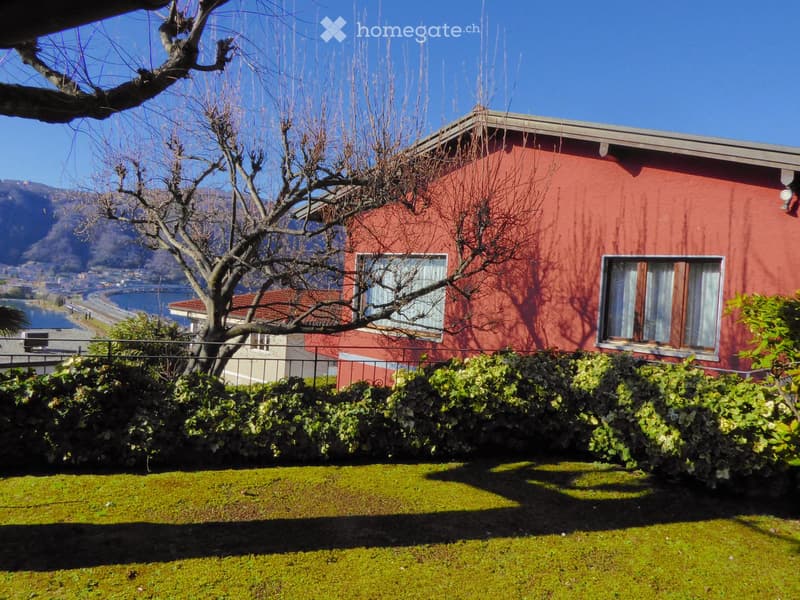 2-story red brick house with large windows, surrounded by a well-maintained garden with lush greenery and a view of the lake or body of water in the background.