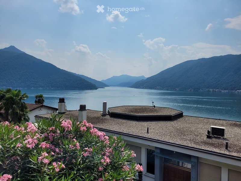 Two-story house with a gravel roof, surrounded by lush greenery and pink flowers. The house overlooks a scenic lake or bay, with mountains in the background. The view from the house appears to be breathtaking.