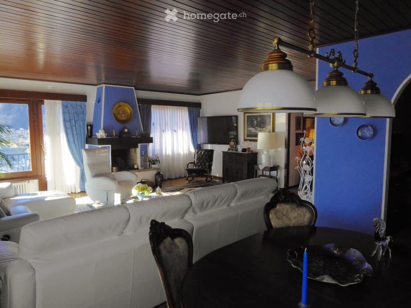 Spacious living room with large windows offering a scenic view, dark wood ceiling, white sofa, antique-style chairs, decorative lamps, and various decorative elements throughout the room.