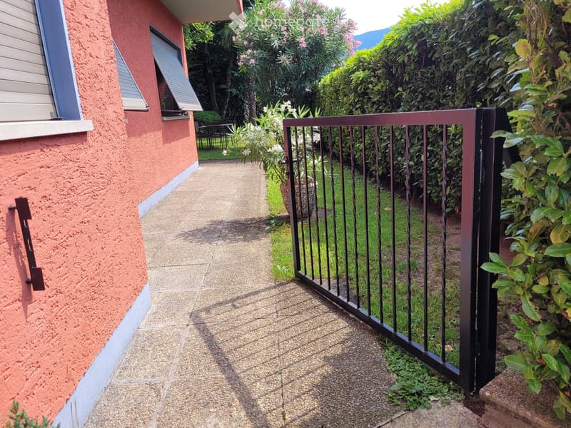 2-story house with red stucco exterior, black metal railing fence, concrete walkway, lush greenery and plants surrounding the property