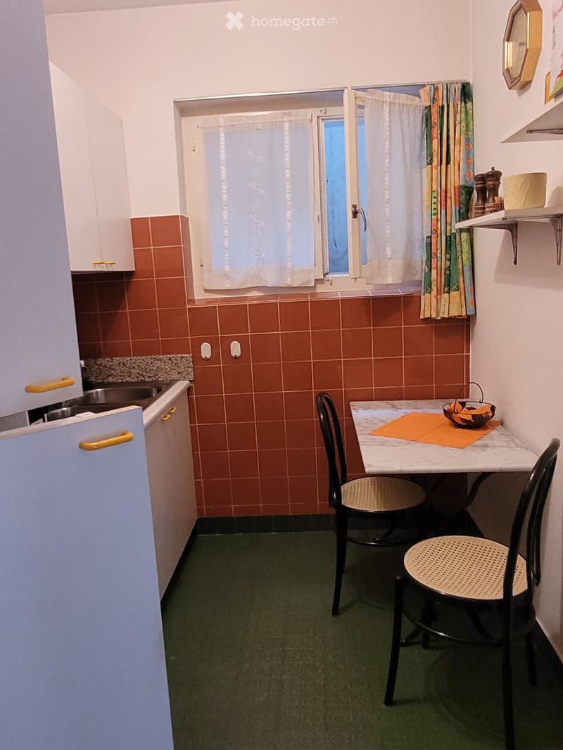 Small kitchen with brown tile walls, a refrigerator, a dining table with two chairs, and a window with curtains.