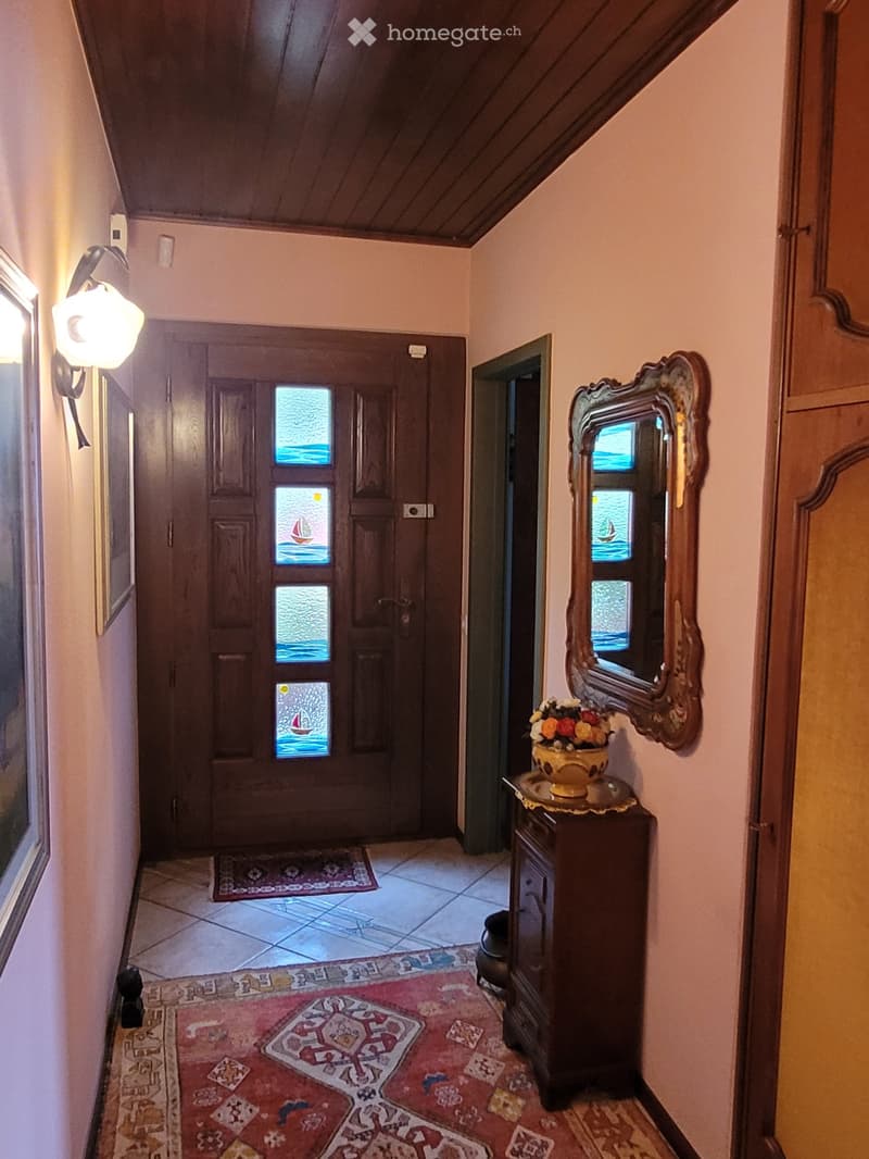 Hallway with wooden ceiling, tiled floor, decorative rugs, wall-mounted light fixture, ornate mirror, small table with flower vase, and stained-glass door.
