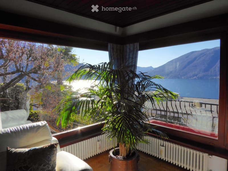 A cozy living space with large windows offering a scenic view of a lake and mountains, featuring a sofa and a potted plant.