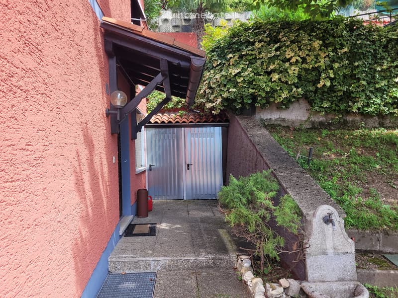 Two-story house with red exterior walls, metal garage door, and lush greenery surrounding the property.