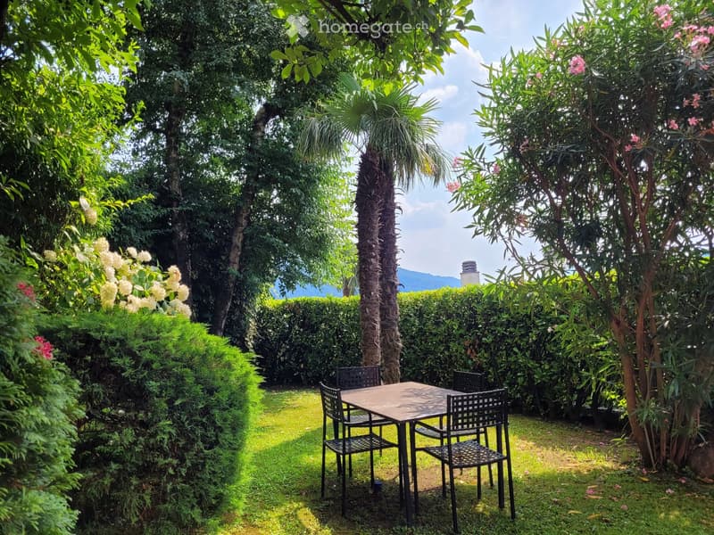 Lush, tropical garden with palm trees, flowering plants, and a patio set with a table and chairs. The garden appears to have a view of the surrounding mountains or hills.