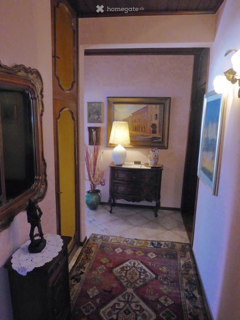 Ornate framed mirror, antique dresser with lamp and decor, patterned area rug, and a fireplace visible in the background.