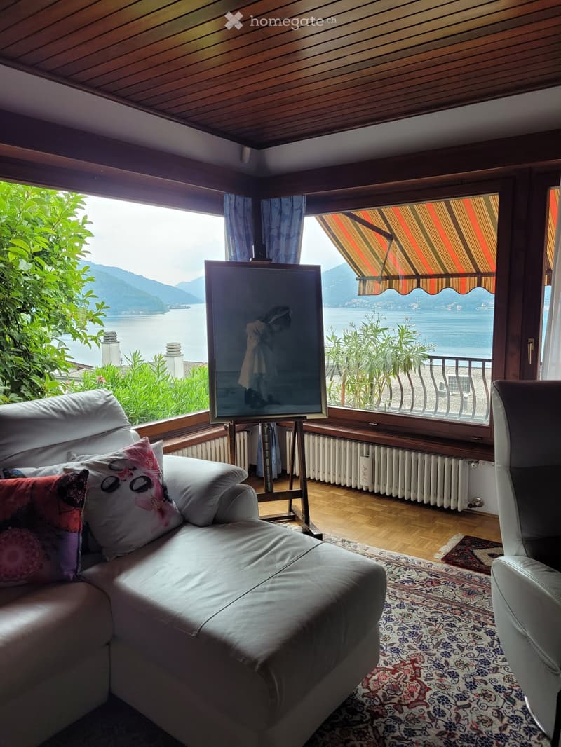Cozy living room with large windows overlooking a scenic lake and mountains, wooden ceiling, colorful patterned rug, leather sofa, and an easel displaying artwork.