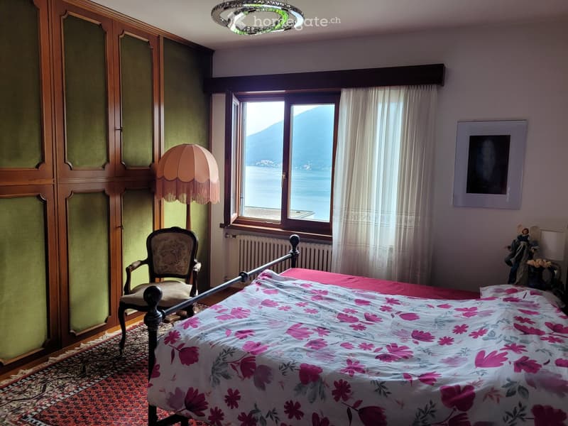 Spacious bedroom with large windows overlooking a lake or body of water, wooden wardrobe, ornate chair, floral bedding, and a decorative ceiling light fixture