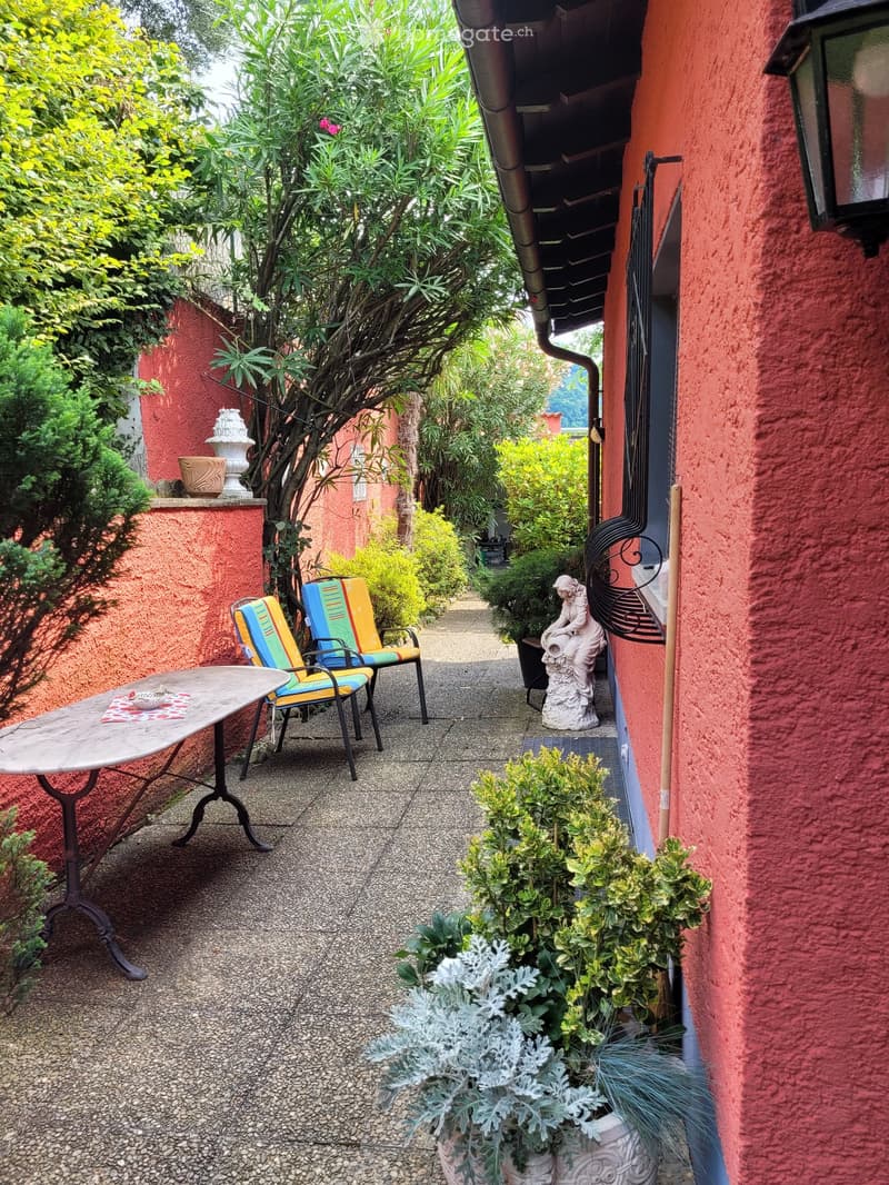 Colorful outdoor patio with red walls, potted plants, a table and chairs, and a decorative fireplace or oven feature. The patio is surrounded by lush greenery and vegetation, creating a cozy and inviting atmosphere.