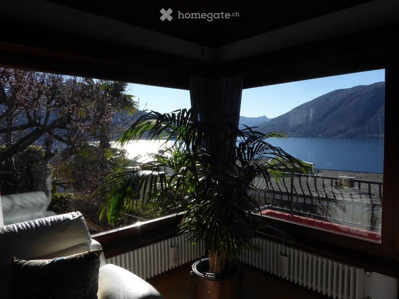 Balcony with a panoramic view of a lake surrounded by mountains. The balcony has a couch and a potted palm tree, providing a relaxing and scenic setting.