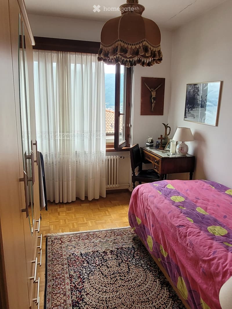 Bedroom with large windows, ornate chandelier, wooden dresser, and a colorful floral bedspread.