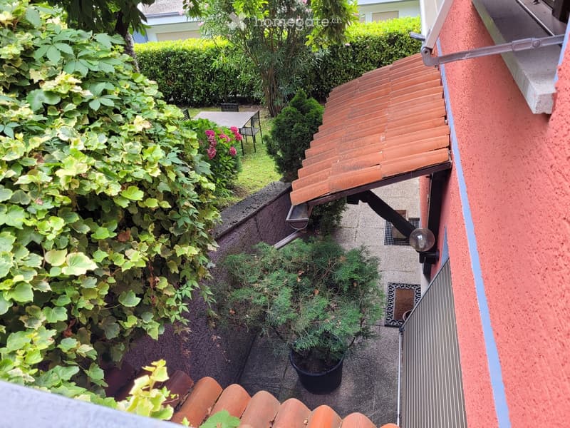 The image shows a well-maintained outdoor garden area with lush greenery, including shrubs, plants, and a small tree. There is a wooden deck or balcony structure with steps leading down to the garden. The garden appears to be surrounded by a brick or ston