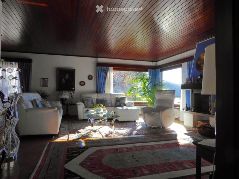 Spacious living room with large windows, wooden paneled ceiling, plush white sofas, glass coffee table, and a potted plant. The room has a cozy and warm atmosphere with natural light streaming in.