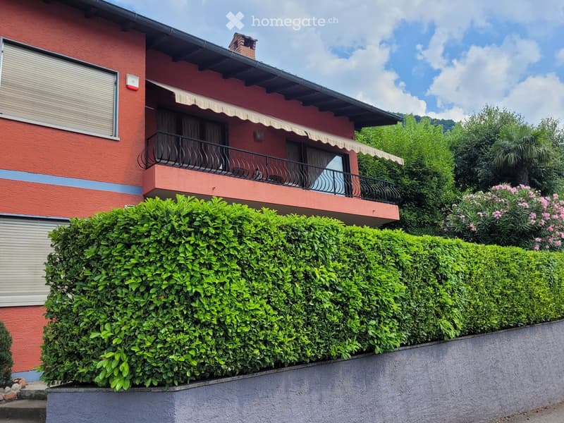 2-story red brick building with a balcony, surrounded by lush green hedges and plants