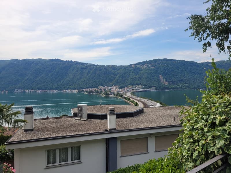 2-story house with a gravel roof, overlooking a lake surrounded by mountains and greenery. The house has large windows and a balcony or terrace with a view of the scenic landscape.