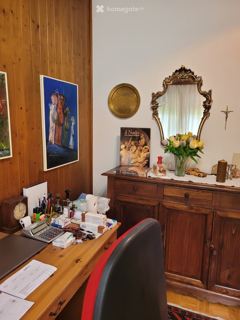Wooden paneled walls, ornate mirror, wooden dresser with decorative items, desk with office supplies, framed artwork on the wall