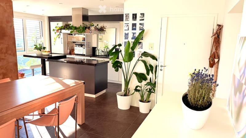 Modern kitchen with a central island, stainless steel appliances, and a dining area with a wooden table and pink chairs. Potted plants are placed around the space, and there is a view of a balcony with outdoor seating.