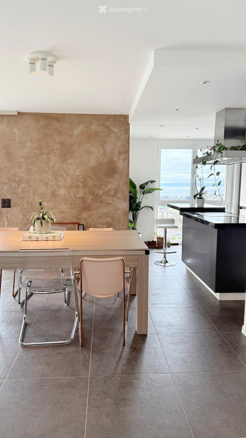 Modern dining area with a wooden table, clear chairs, a stool, and a counter, featuring a view of the ocean through large windows.