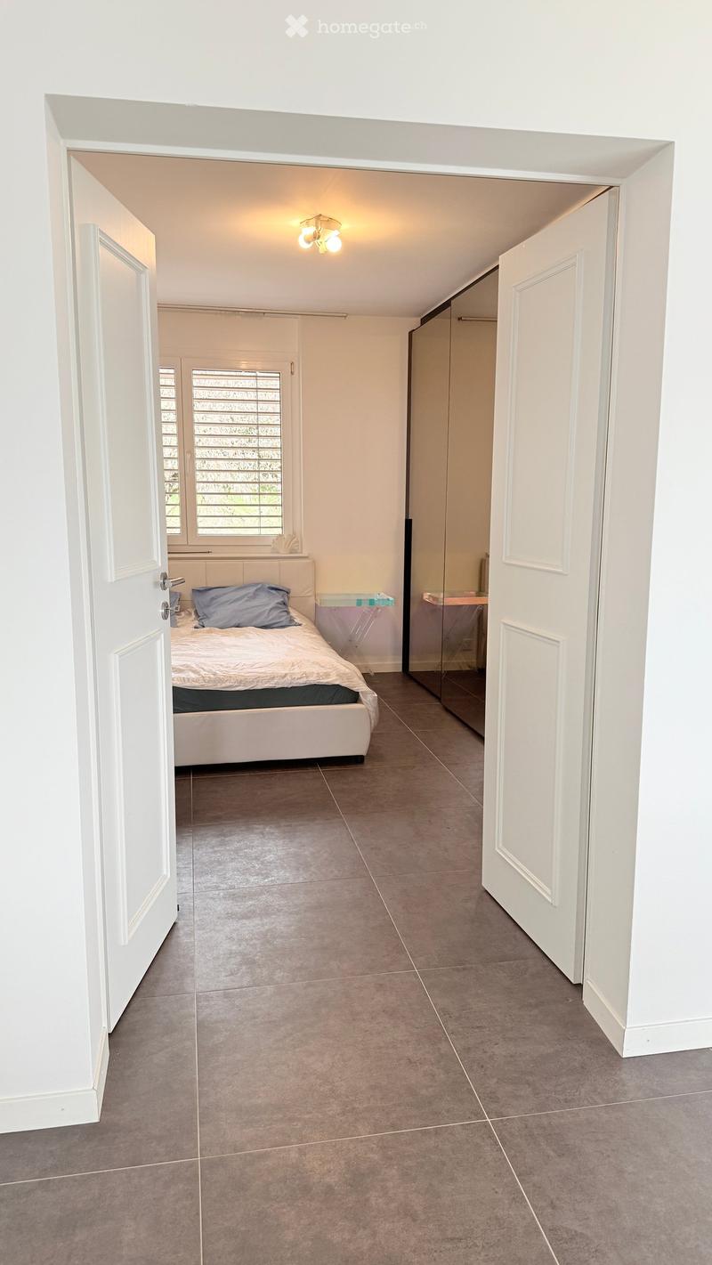 A clean, modern hallway with tiled flooring, white walls, and a ceiling light fixture. The hallway leads to a bedroom with a bed, pillows, and a small table with a lamp.