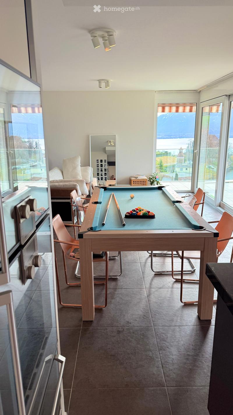 A modern living room with a pool table, surrounded by chairs and a sofa, large windows with scenic views, and a mirror on the wall.