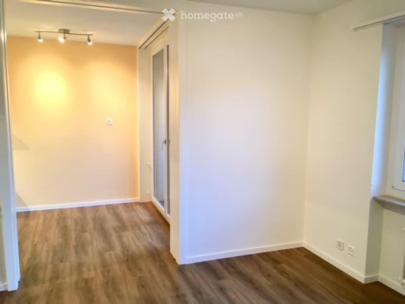Empty room with light wooden flooring and white walls, featuring a small window and a door leading to another room.